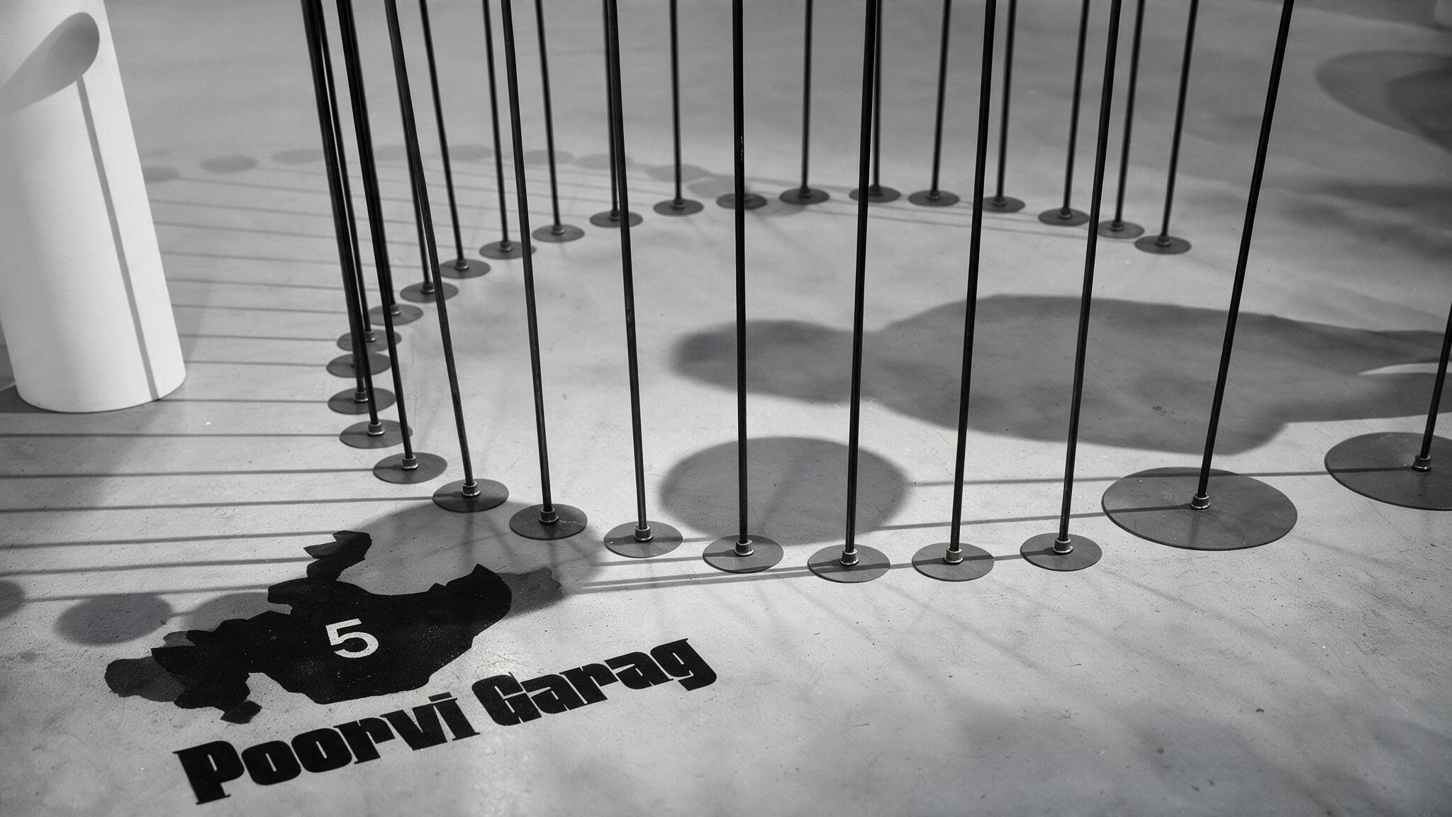An overhead view of a spiral installation of pins and rods on a staircase, casting a shadow shaped like a number 5 with the text 'Poorvi Garag'