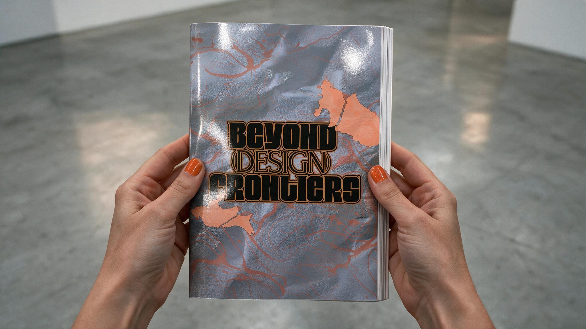 Two hands holding a marbled 'Beyond Design Frontiers' booklet over a concrete floor