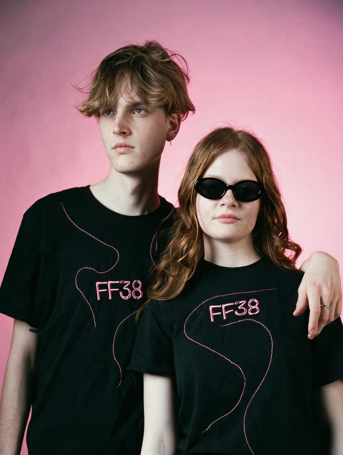 A young man and woman in sunglasses, both wearing black FF38 t-shirts with embroidered pink squiggle designs, against a pink backdrop