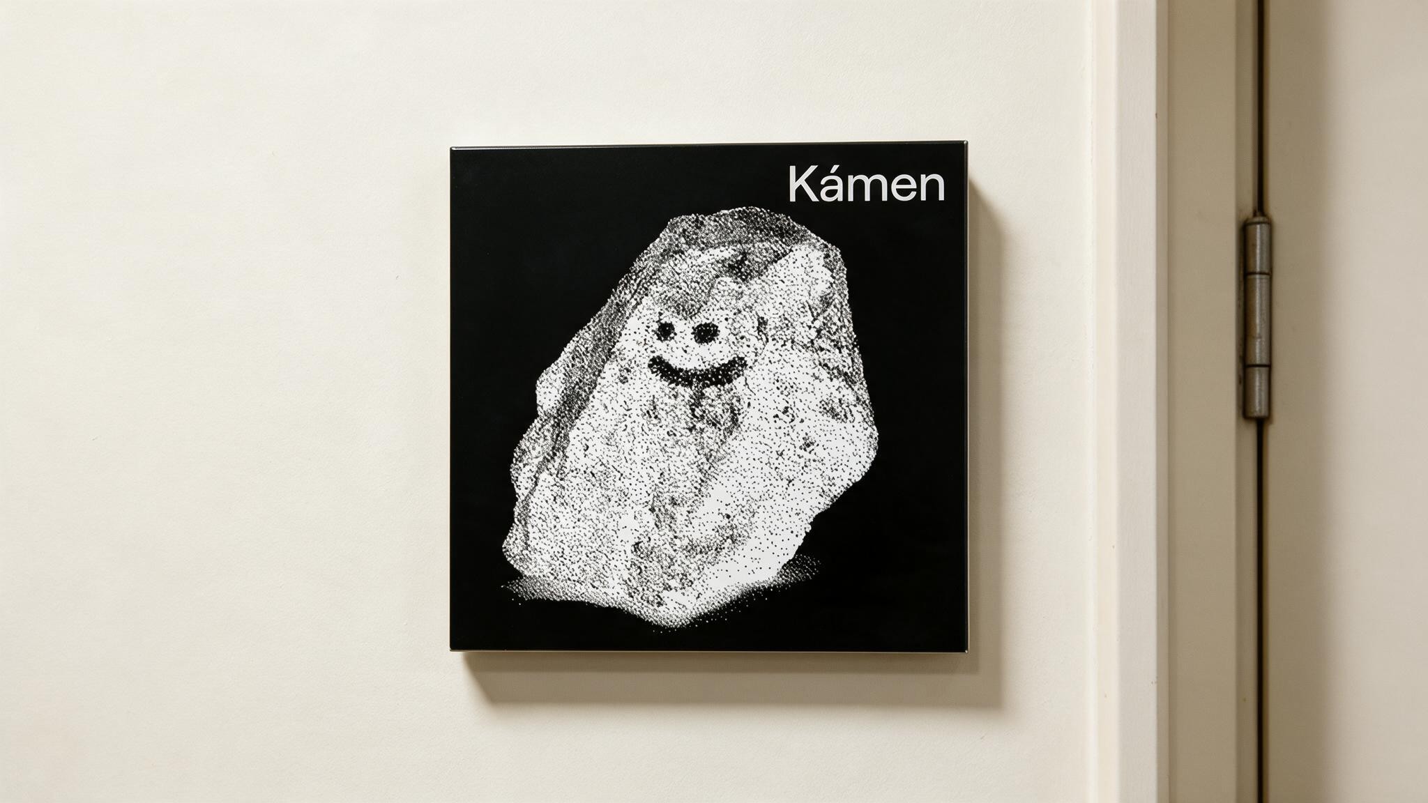 A black door sign reading 'Kámen' illustrated with a smiling stippled rock, mounted beside a door