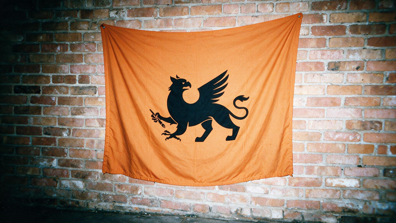 An orange flag printed with the Kuja griffin mark, hung against a brick wall