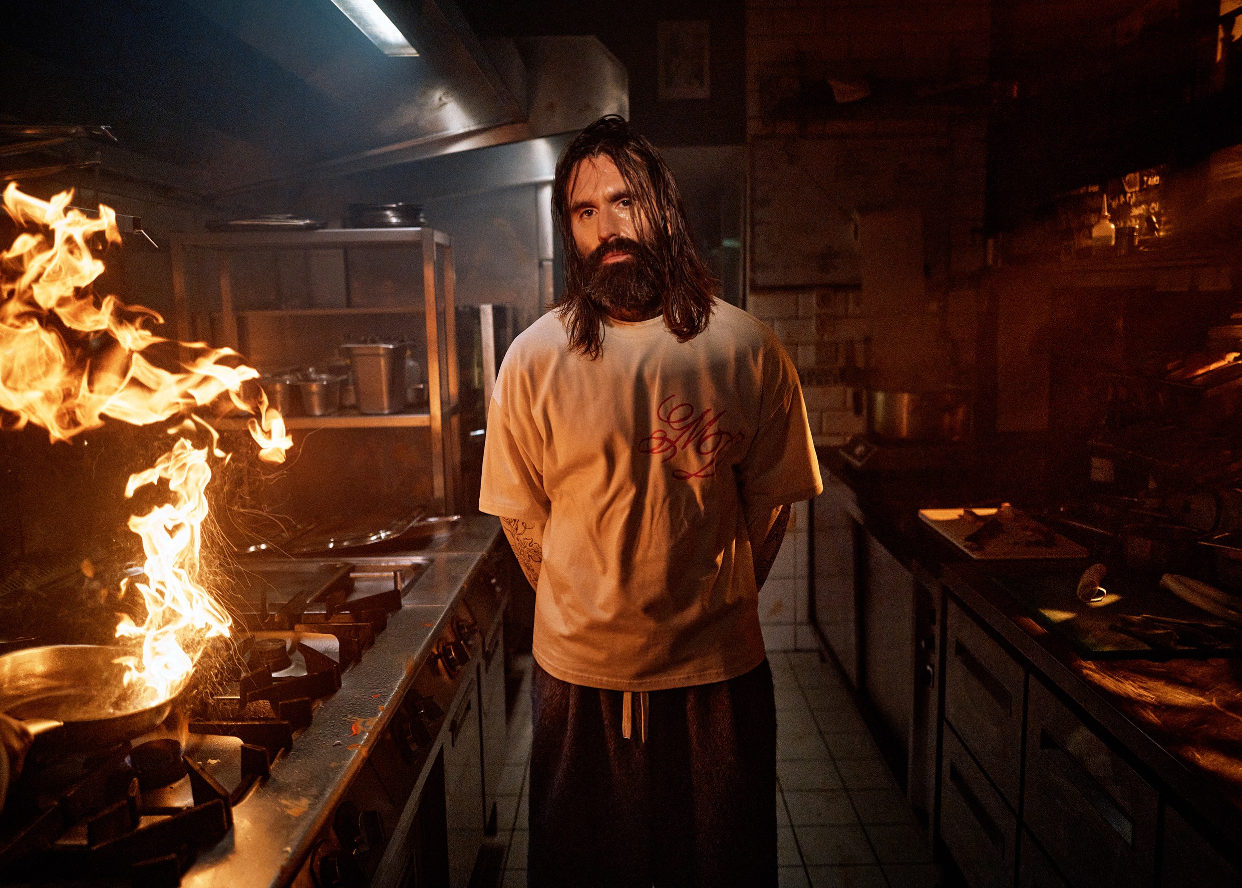 A bearded chef in a cream My Dear t-shirt stands in a restaurant kitchen beside open flame