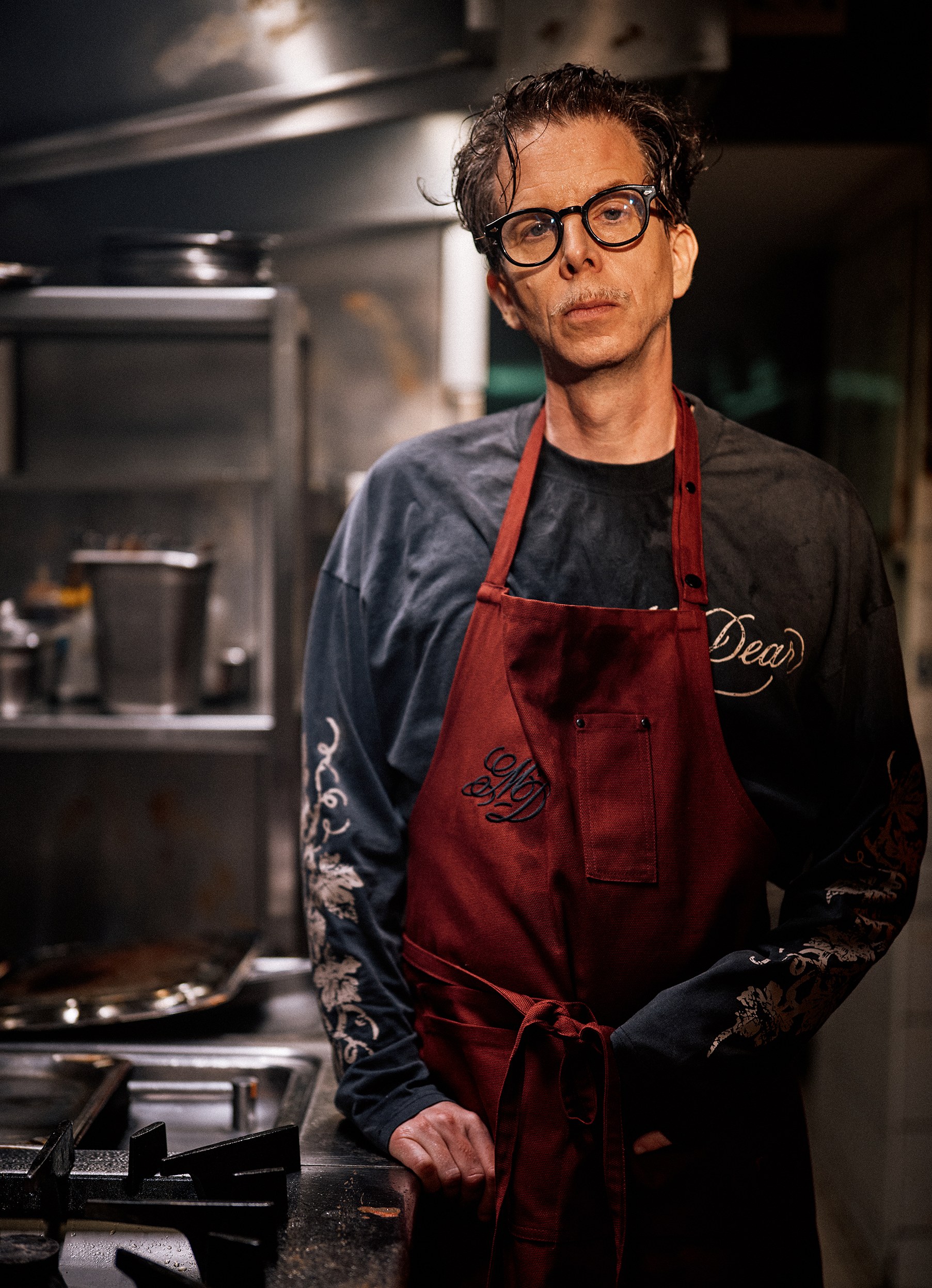 A chef wearing glasses and a red My Dear apron over a long-sleeve top printed with floral tattoo graphics