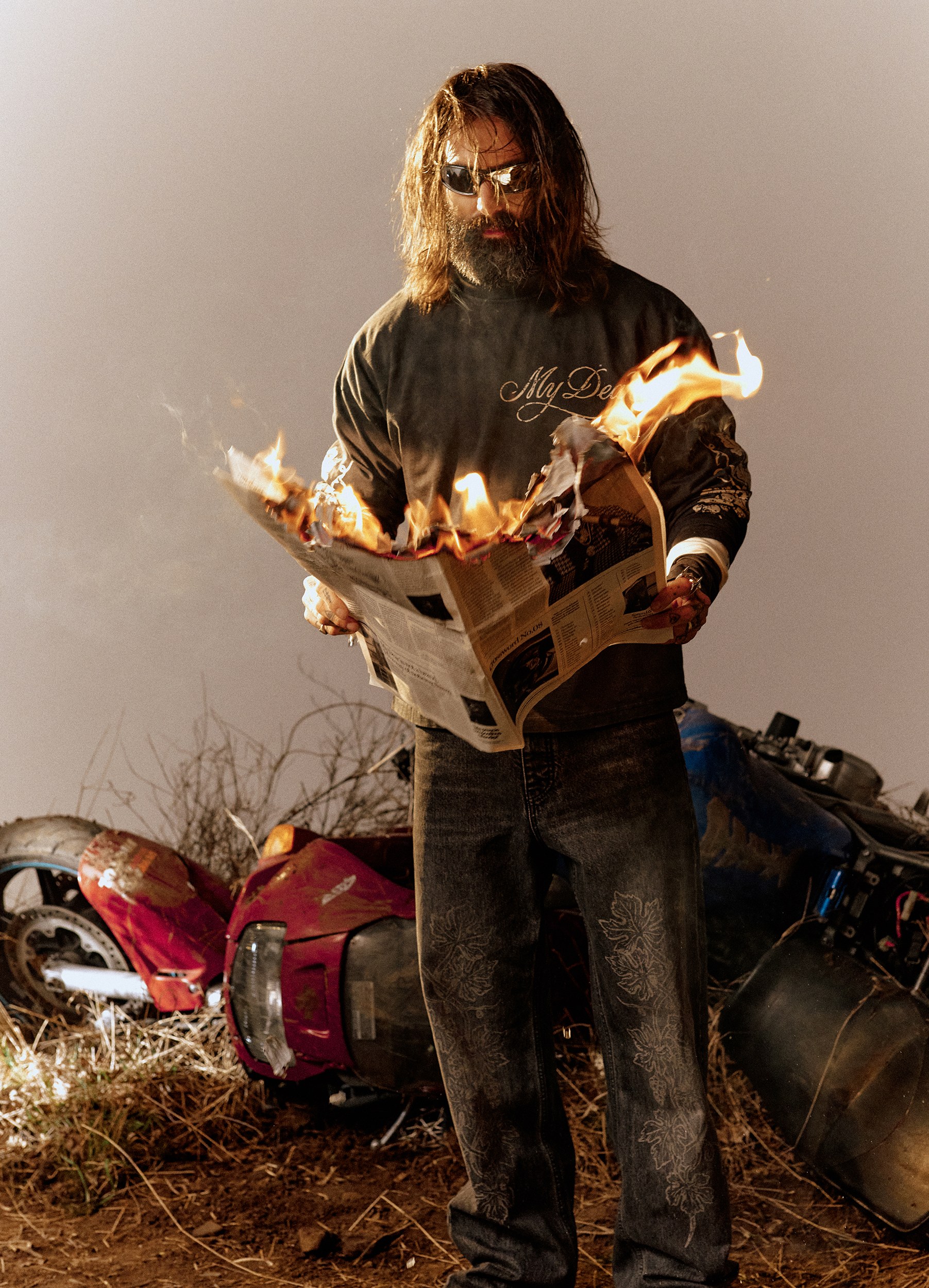 A man in sunglasses and a My Dear sweater burning a newspaper outdoors beside wrecked motorcycles