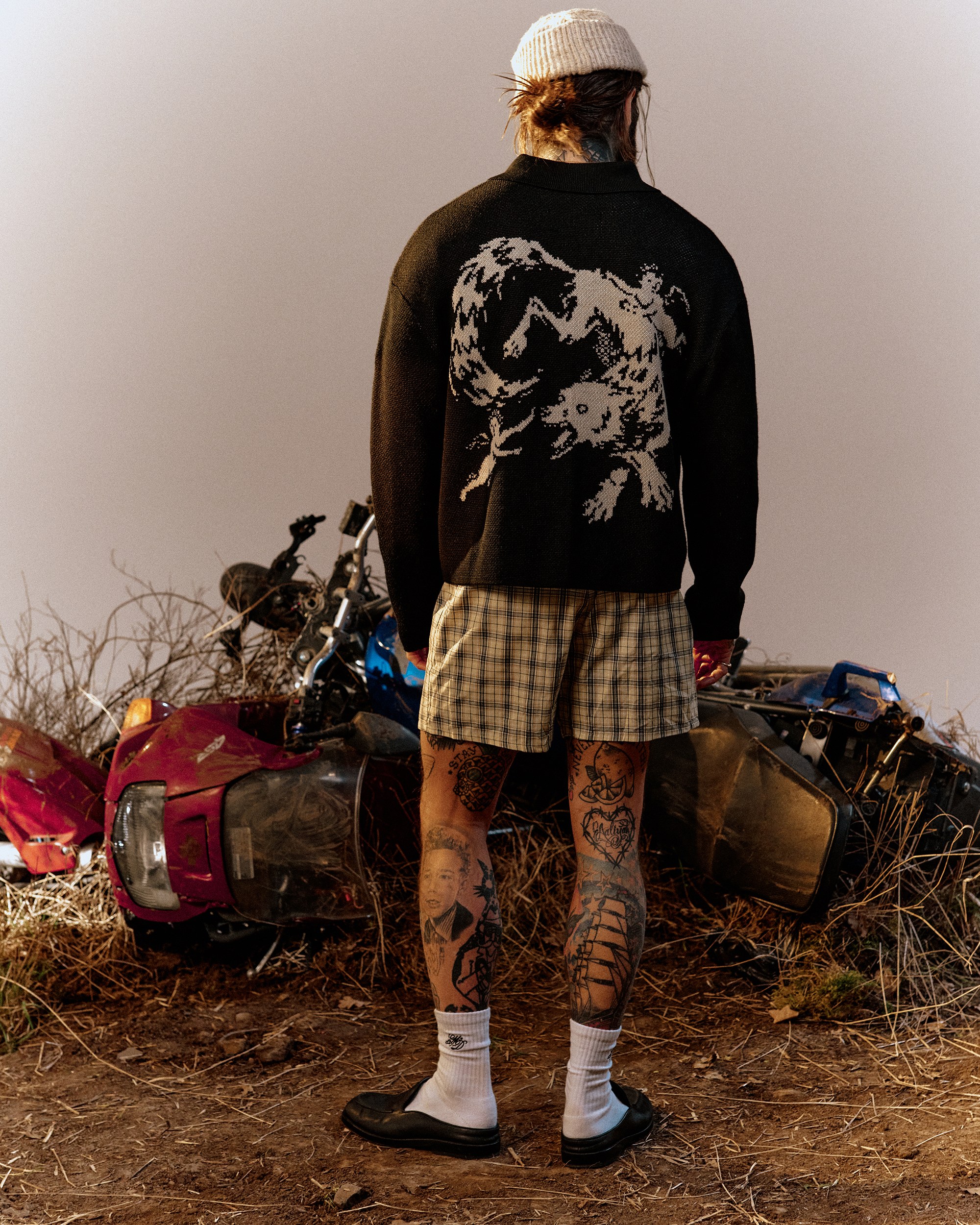 A man seen from behind wearing a black sweater with a skeleton cow knit graphic, standing before wrecked vehicles