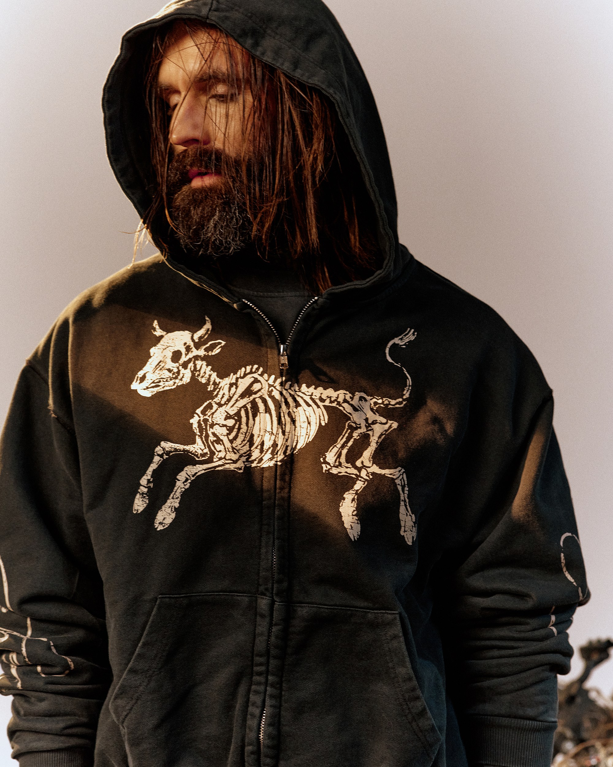 Close-up of a hooded man wearing a My Dear zip hoodie printed with a skeleton cow graphic