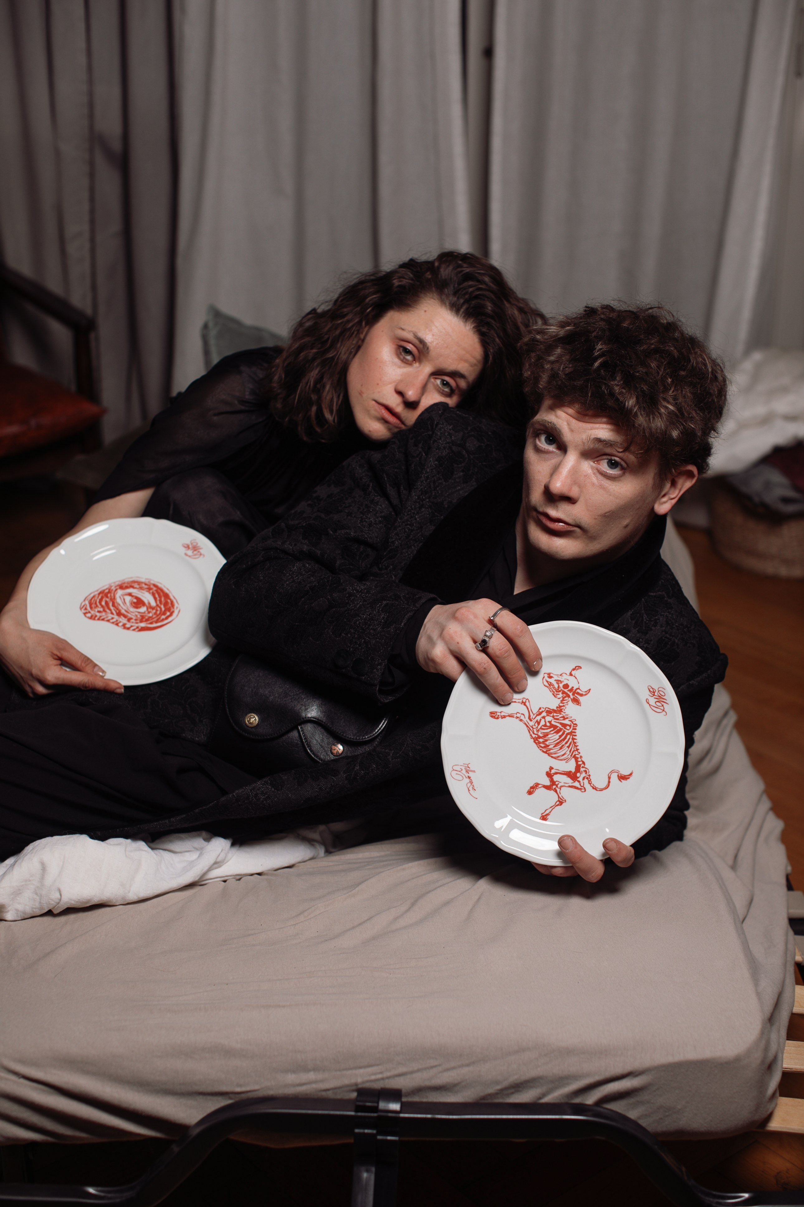 Two people lying on a bed, each holding a porcelain plate hand-painted with red skeleton motifs