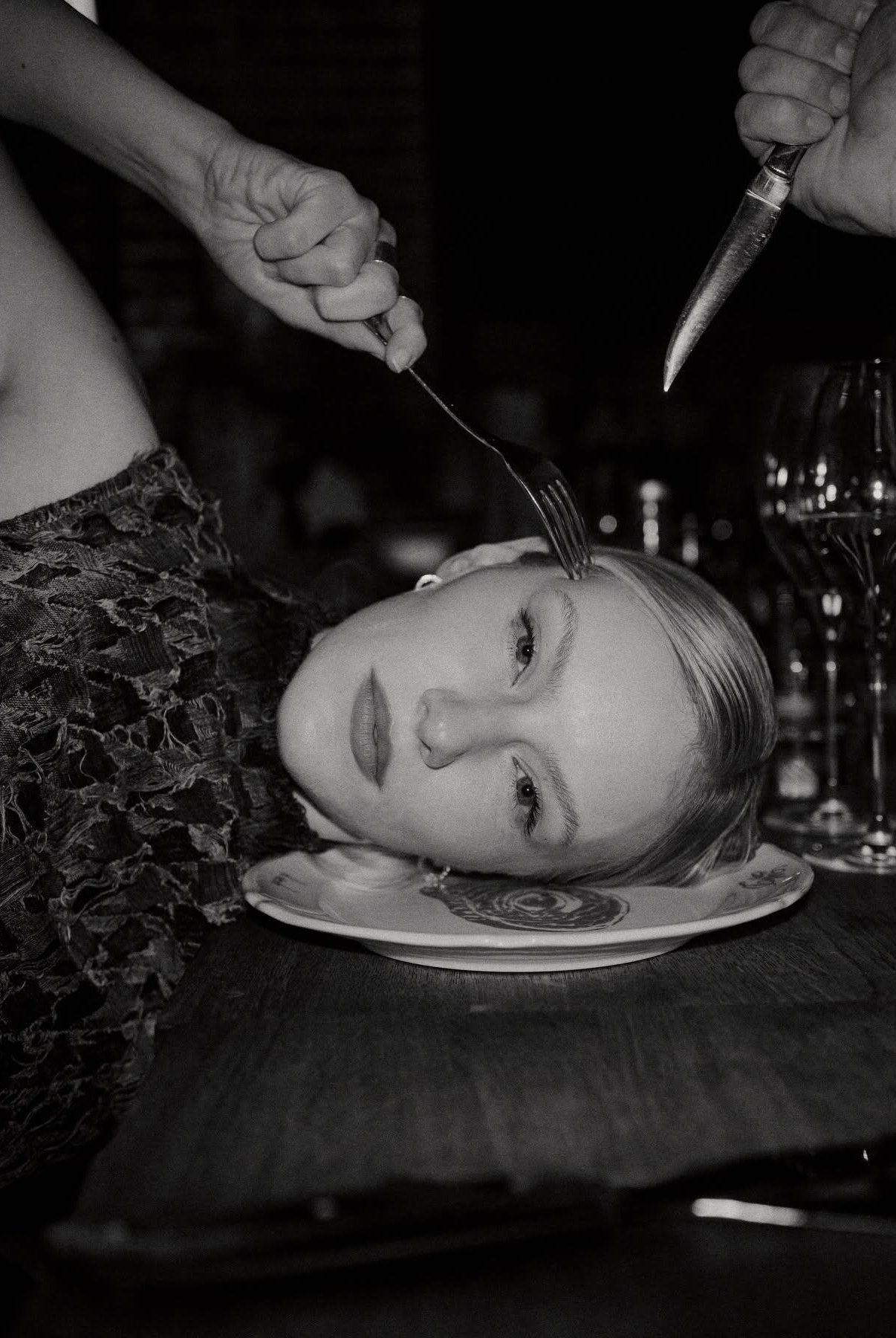 Black and white photo of hands plating a sculptural head-shaped dish with a fork and knife at a restaurant table