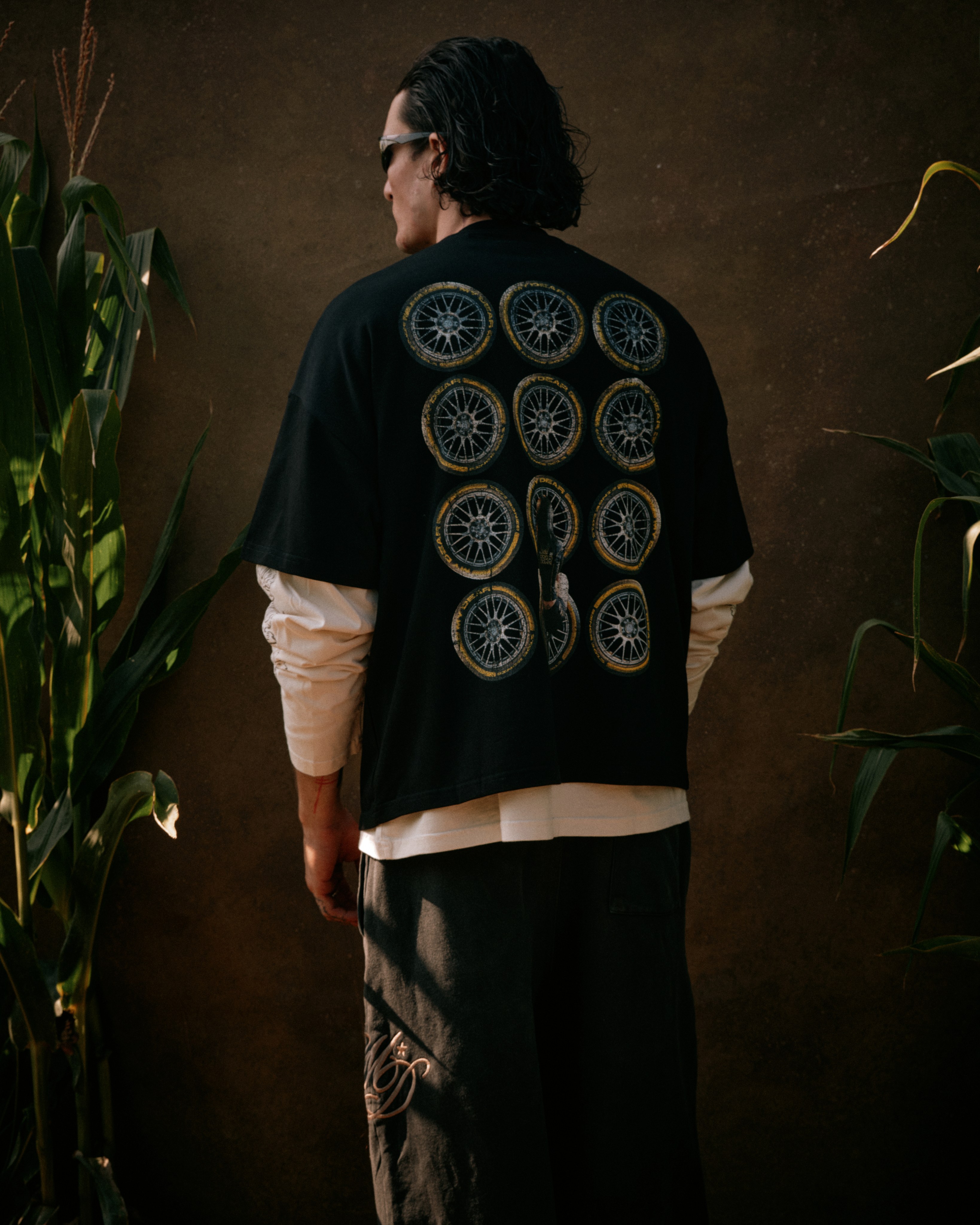 A man seen from behind wearing a black t-shirt printed with a grid of car wheel graphics, standing in a cornfield