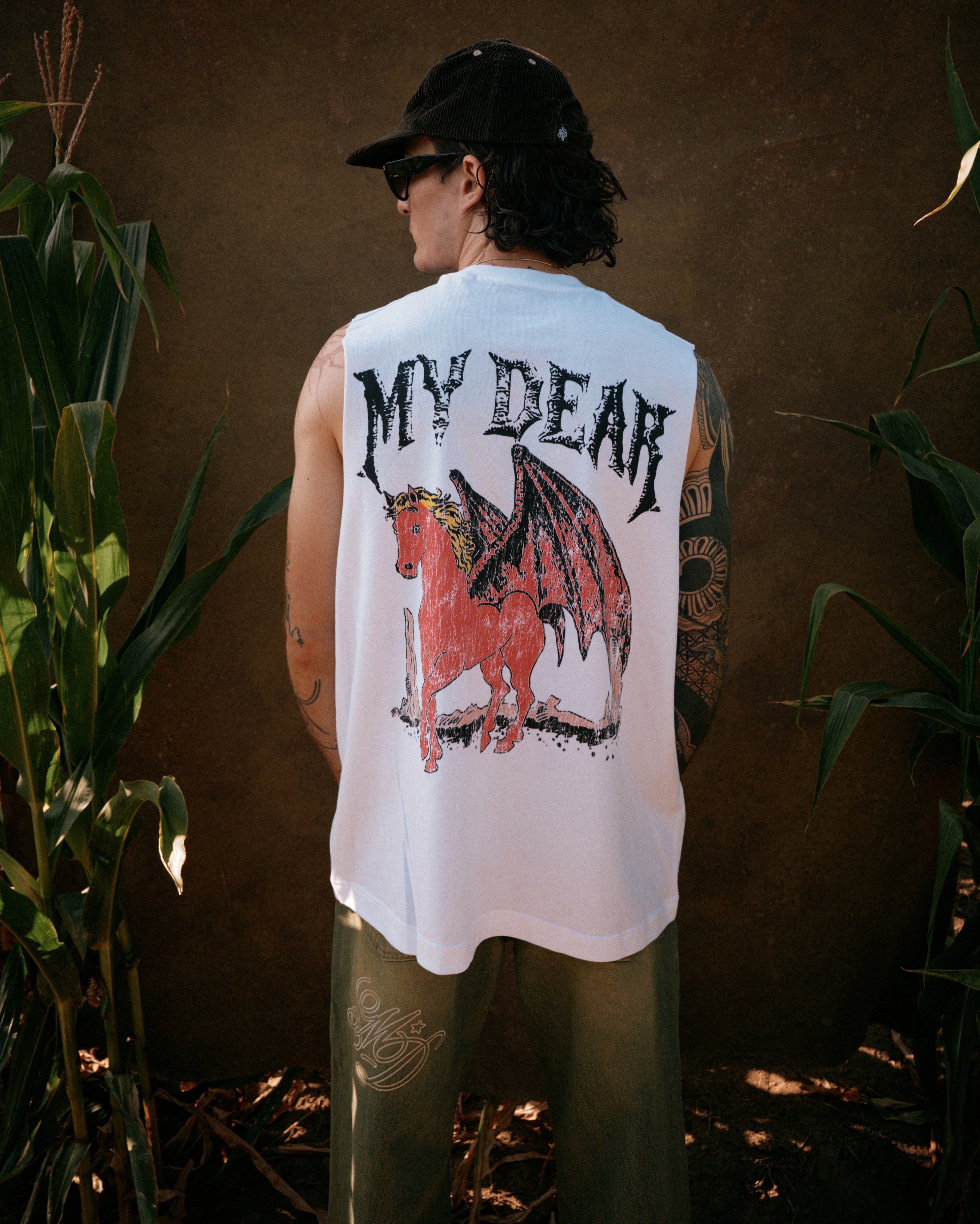 A man seen from behind wearing a white sleeveless My Dear shirt printed with a winged, horned horse graphic, standing in a cornfield