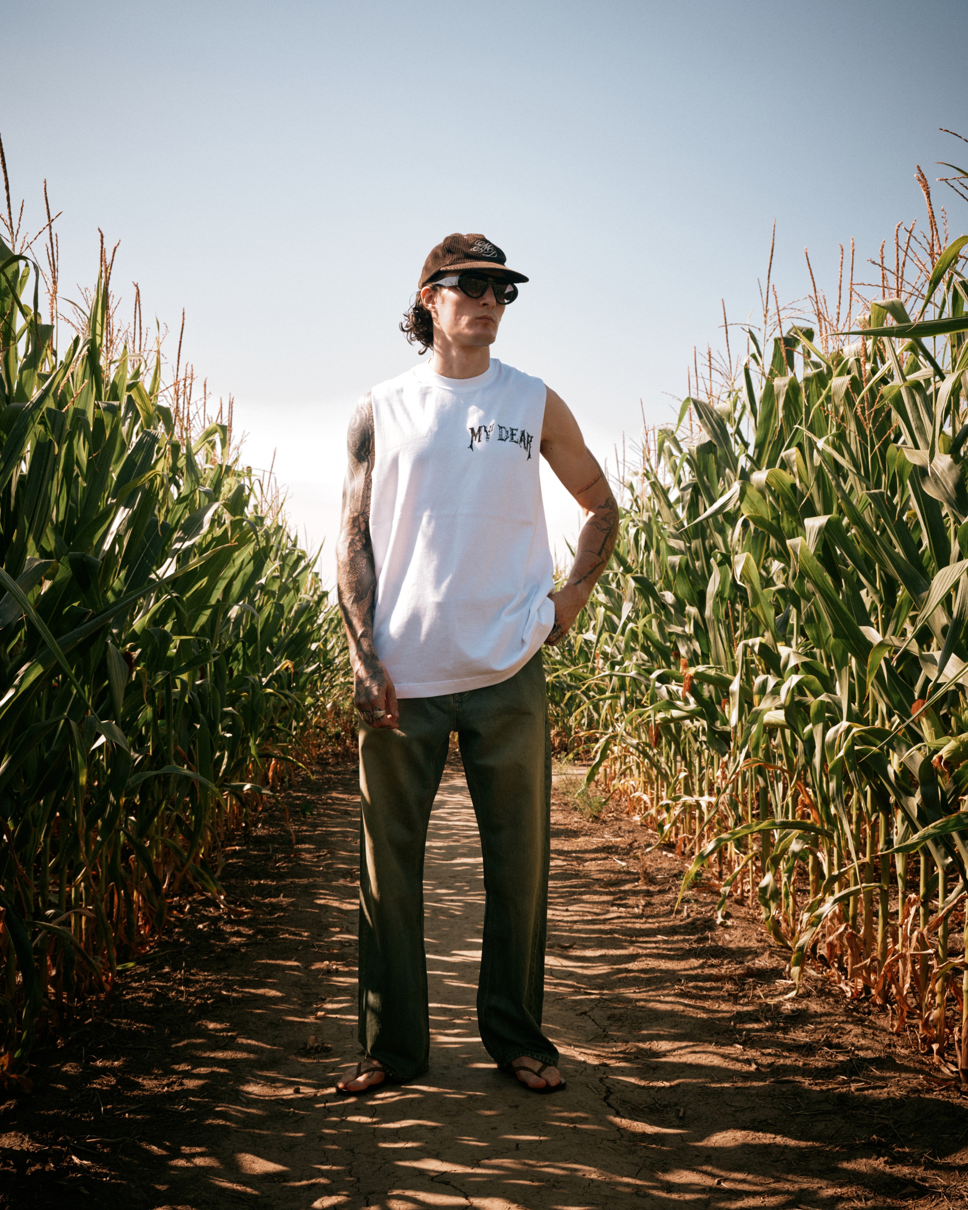 A man in sunglasses and a white My Dear tank top standing on a dirt path between rows of corn