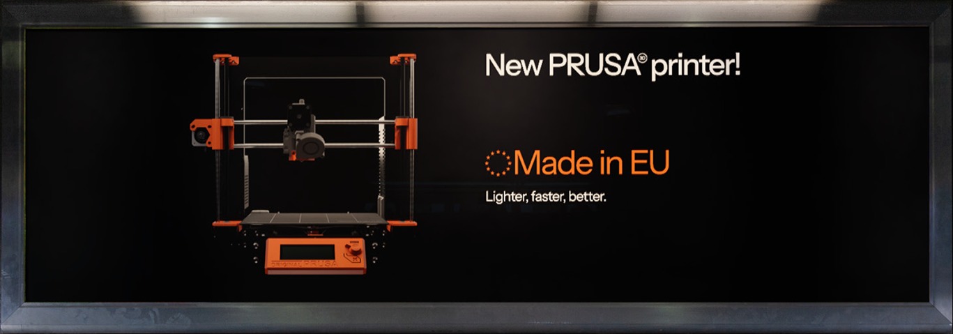 A wall-mounted monitor ad reading 'New PRUSA printer! Made in EU. Lighter, faster, better.' beside a render of the printer